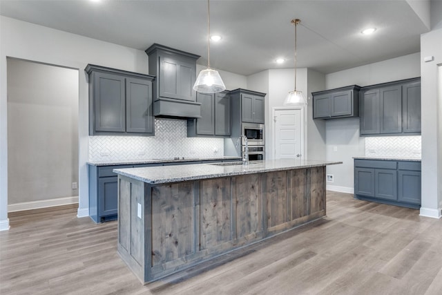 kitchen with appliances with stainless steel finishes, a kitchen island with sink, hanging light fixtures, light stone counters, and extractor fan