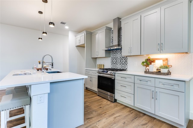 kitchen with sink, stainless steel gas stove, an island with sink, decorative light fixtures, and wall chimney exhaust hood