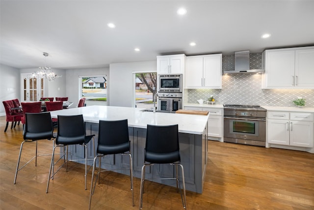 kitchen featuring hanging light fixtures, appliances with stainless steel finishes, wall chimney range hood, a kitchen island with sink, and white cabinets