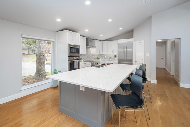 kitchen featuring built in appliances, light stone counters, white cabinets, a center island with sink, and wall chimney exhaust hood