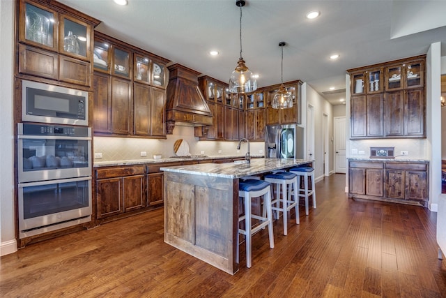 kitchen featuring a kitchen island with sink, stainless steel appliances, light stone counters, decorative light fixtures, and custom exhaust hood