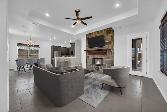 living area featuring visible vents, baseboards, a tray ceiling, a stone fireplace, and ceiling fan with notable chandelier