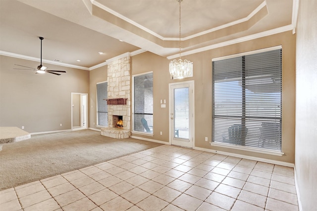 unfurnished living room with ceiling fan with notable chandelier, a tray ceiling, ornamental molding, a stone fireplace, and light colored carpet