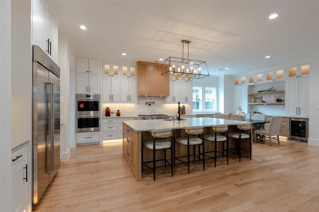 kitchen with light hardwood / wood-style flooring, appliances with stainless steel finishes, an island with sink, beverage cooler, and white cabinets