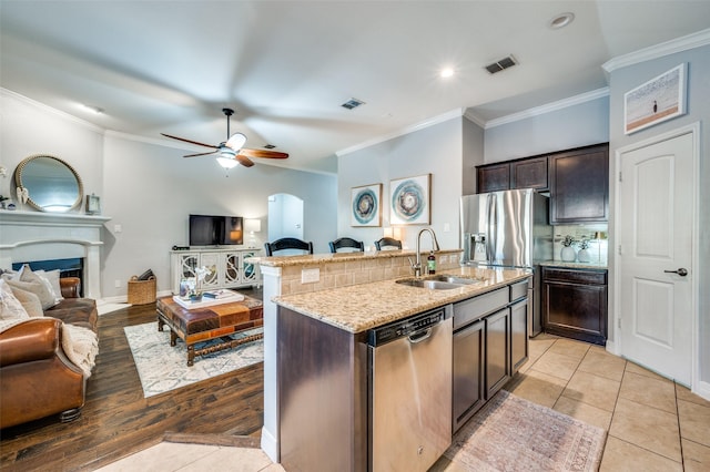 kitchen with dark brown cabinetry, sink, ornamental molding, stainless steel appliances, and a kitchen island with sink