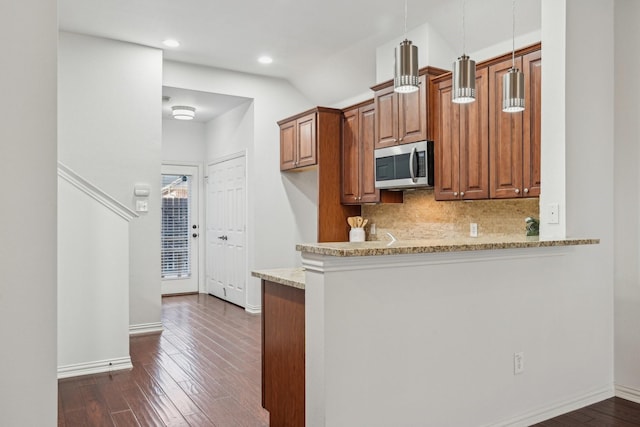 kitchen featuring light stone counters, dark wood-style floors, stainless steel microwave, hanging light fixtures, and brown cabinetry