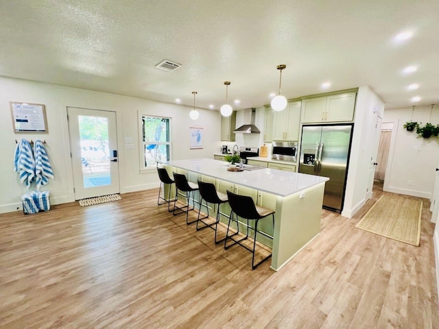 kitchen featuring wall chimney range hood, a breakfast bar, appliances with stainless steel finishes, a kitchen island with sink, and hanging light fixtures