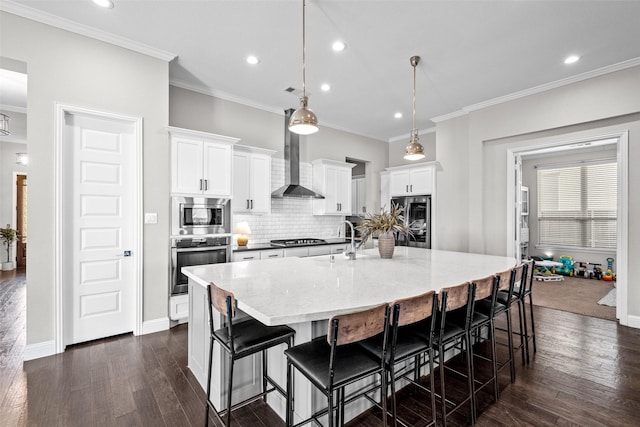 kitchen with ornamental molding, a large island with sink, appliances with stainless steel finishes, wall chimney exhaust hood, and dark wood-style flooring