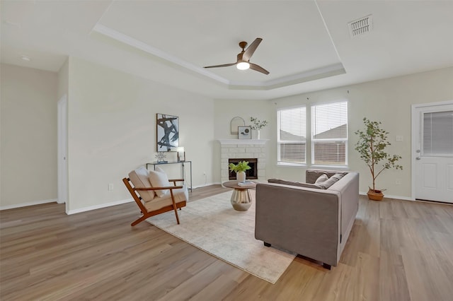 living room featuring ceiling fan, a tray ceiling, a brick fireplace, and light hardwood / wood-style flooring