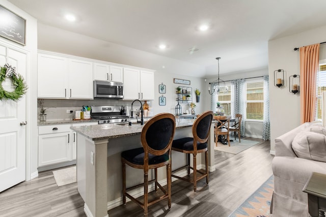 kitchen with appliances with stainless steel finishes, hanging light fixtures, light stone counters, white cabinets, and a center island with sink