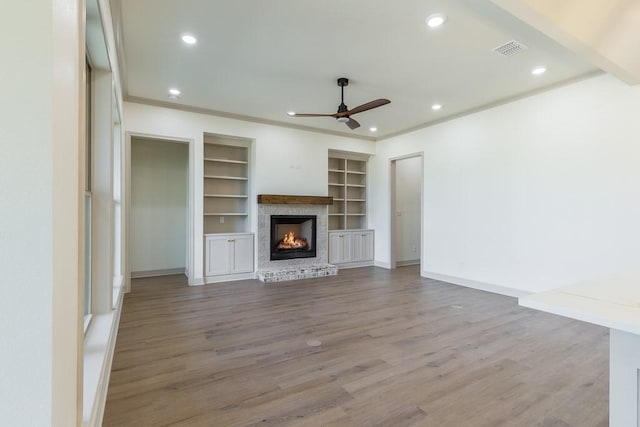 unfurnished living room featuring a fireplace, wood-type flooring, ceiling fan, crown molding, and built in shelves