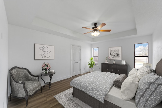 bedroom with multiple windows, dark wood-type flooring, ceiling fan, and a tray ceiling