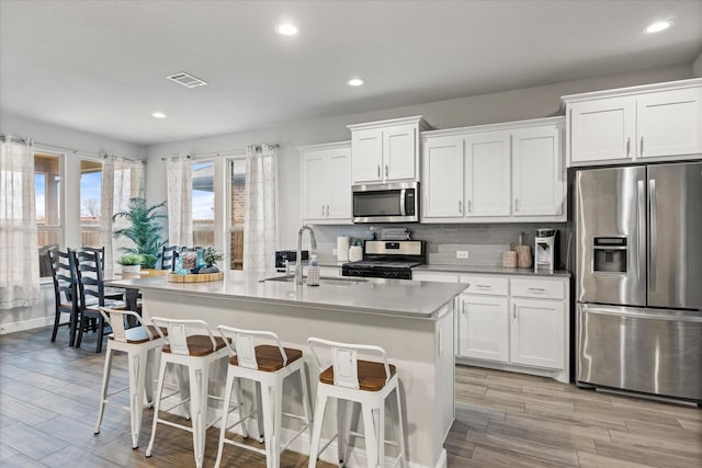 kitchen with sink, a kitchen island with sink, stainless steel appliances, tasteful backsplash, and white cabinets