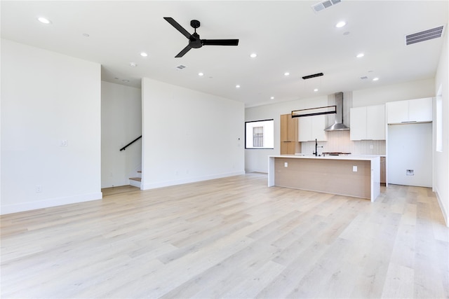 kitchen with wall chimney range hood, white cabinetry, hanging light fixtures, a center island with sink, and light wood-type flooring