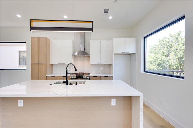 kitchen with wall chimney exhaust hood, white cabinetry, a center island with sink, light stone countertops, and backsplash