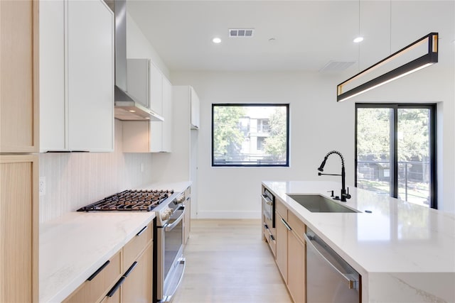 kitchen with sink, wall chimney range hood, appliances with stainless steel finishes, light stone countertops, and a center island with sink