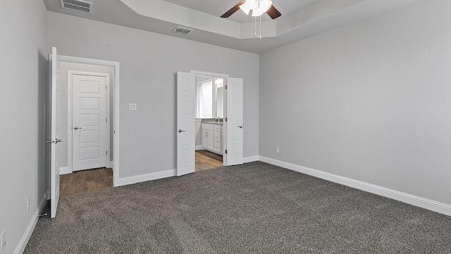 unfurnished bedroom featuring ceiling fan, ensuite bathroom, a tray ceiling, and dark colored carpet