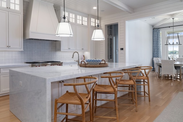 kitchen with white cabinetry, decorative light fixtures, custom exhaust hood, and an island with sink