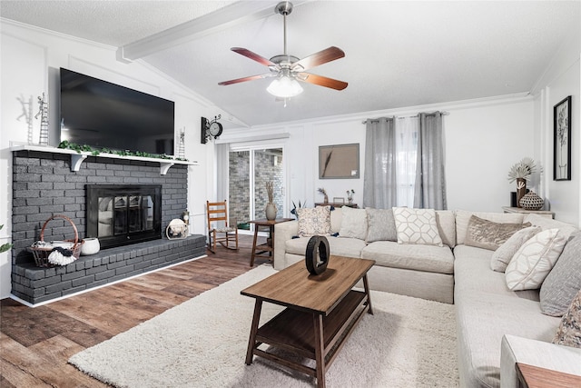 living room featuring wood-type flooring, vaulted ceiling with beams, ornamental molding, ceiling fan, and a brick fireplace