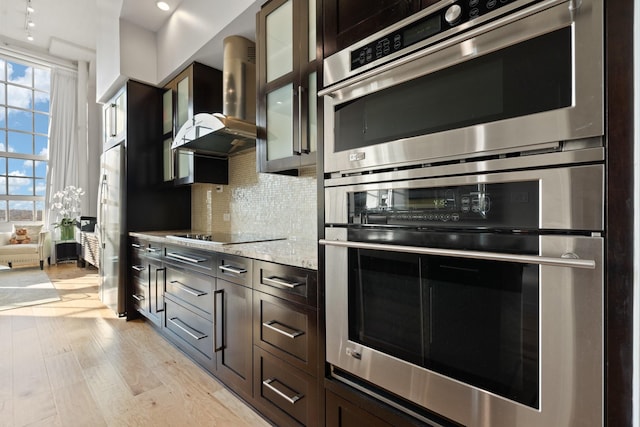 kitchen with tasteful backsplash, light wood-type flooring, stainless steel appliances, light stone countertops, and wall chimney range hood