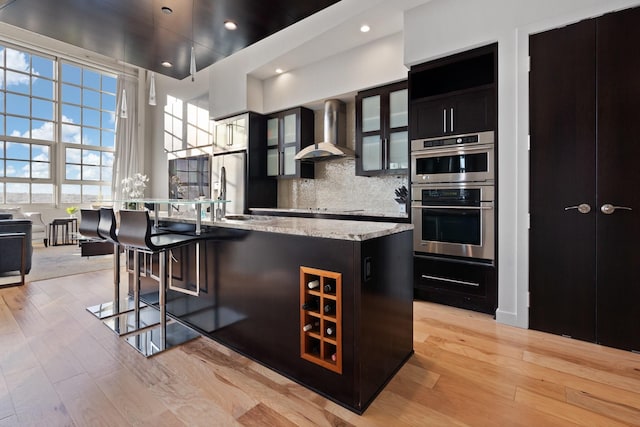 kitchen with a breakfast bar area, double oven, an island with sink, decorative backsplash, and wall chimney exhaust hood