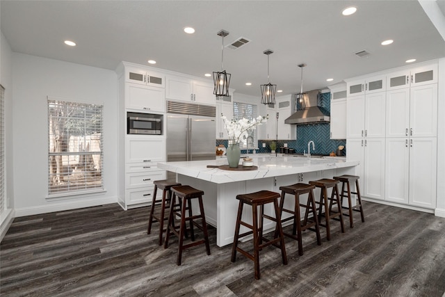 kitchen with a large island with sink, hanging light fixtures, wall chimney range hood, and built in appliances