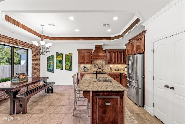 kitchen with premium range hood, pendant lighting, sink, stainless steel fridge, and a center island with sink