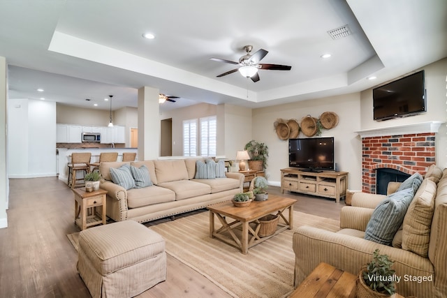 living room featuring a raised ceiling, ceiling fan, a fireplace, and light hardwood / wood-style flooring