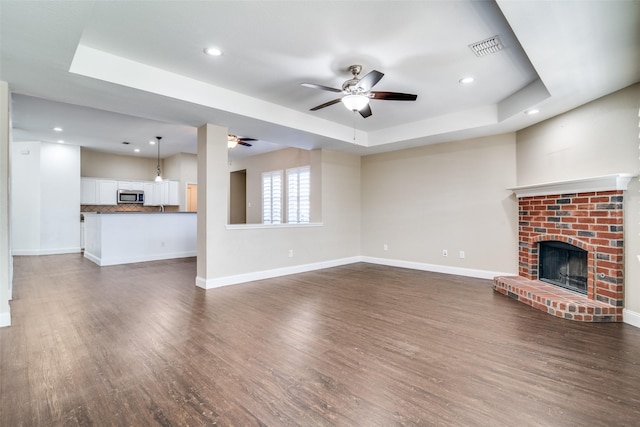 unfurnished living room featuring dark wood-type flooring, ceiling fan, a tray ceiling, and a brick fireplace