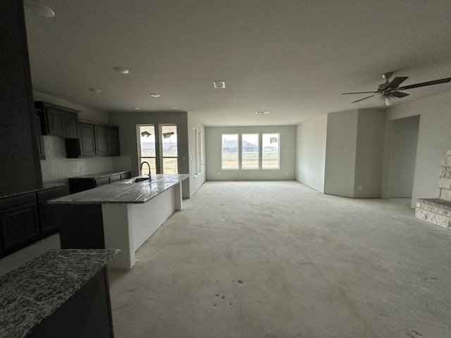 kitchen with an island with sink, sink, ceiling fan, and dark stone counters