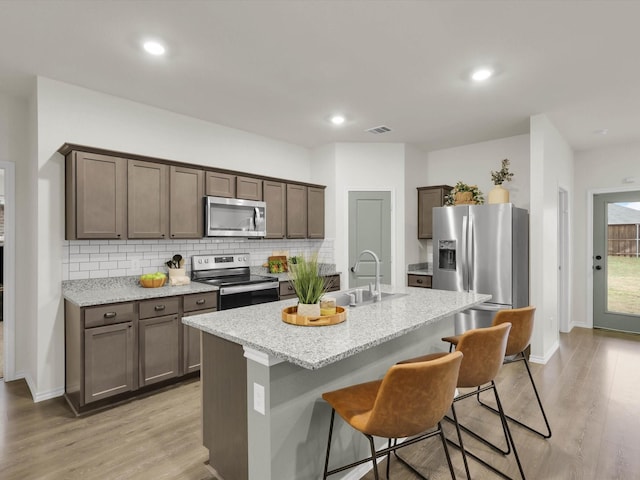 kitchen with stainless steel appliances, tasteful backsplash, light stone counters, an island with sink, and light wood-type flooring