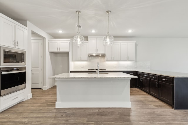 kitchen with built in microwave, white cabinetry, stainless steel oven, hanging light fixtures, and a kitchen island with sink