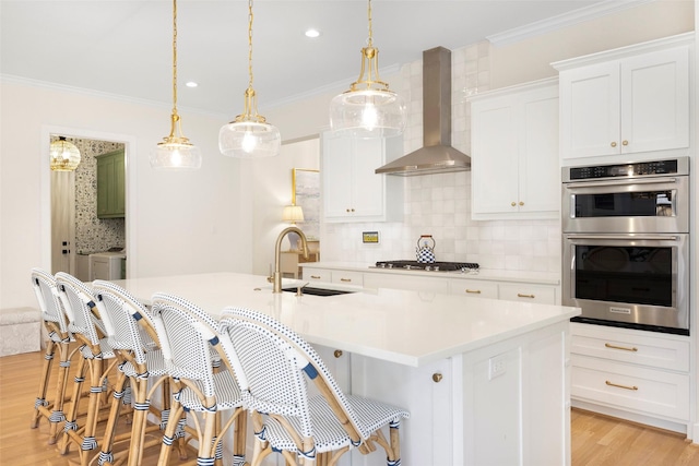 kitchen with appliances with stainless steel finishes, white cabinetry, sink, a kitchen island with sink, and wall chimney exhaust hood