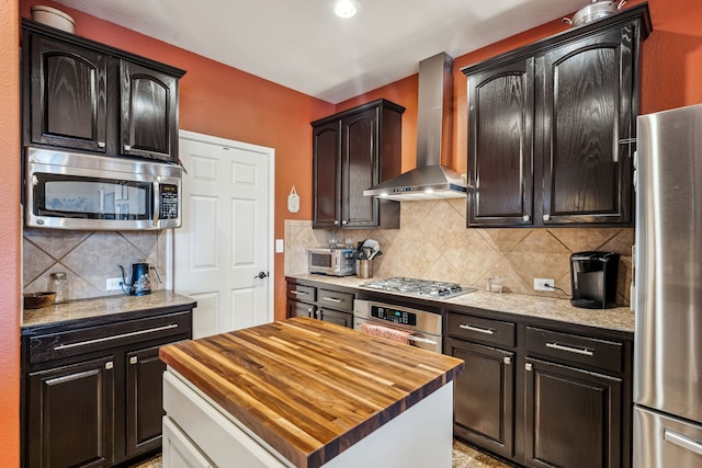 kitchen with appliances with stainless steel finishes, dark brown cabinetry, a kitchen island, wood counters, and wall chimney exhaust hood