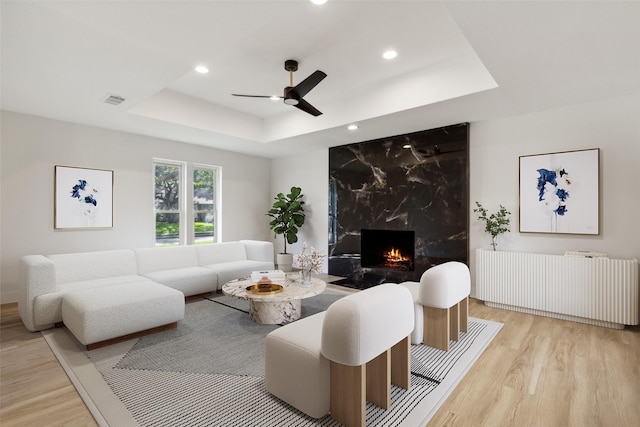 living room with a tray ceiling, radiator heating unit, ceiling fan, light hardwood / wood-style floors, and a premium fireplace