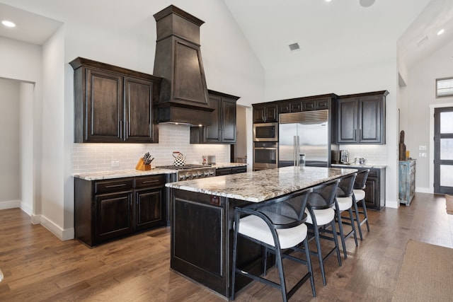 kitchen with built in appliances, a breakfast bar area, premium range hood, light stone countertops, and dark wood finished floors