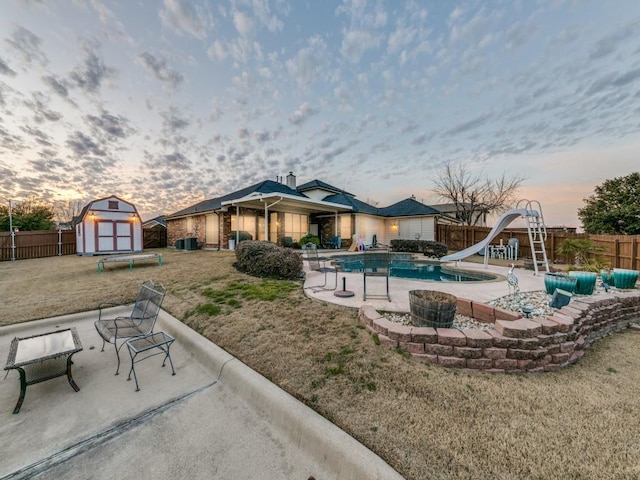 pool at dusk with a storage shed, a patio area, a lawn, and a water slide