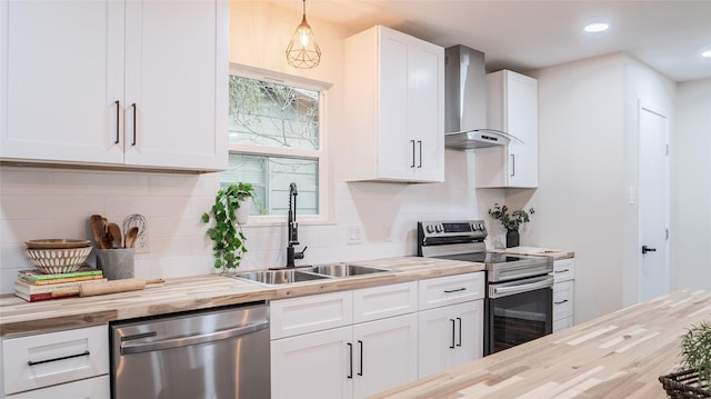 kitchen featuring butcher block countertops, white cabinetry, hanging light fixtures, stainless steel appliances, and wall chimney range hood