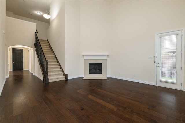 unfurnished living room featuring arched walkways, dark wood finished floors, a tile fireplace, a towering ceiling, and stairs