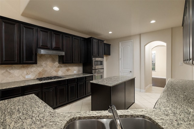 kitchen featuring light stone counters, arched walkways, tasteful backsplash, appliances with stainless steel finishes, and under cabinet range hood