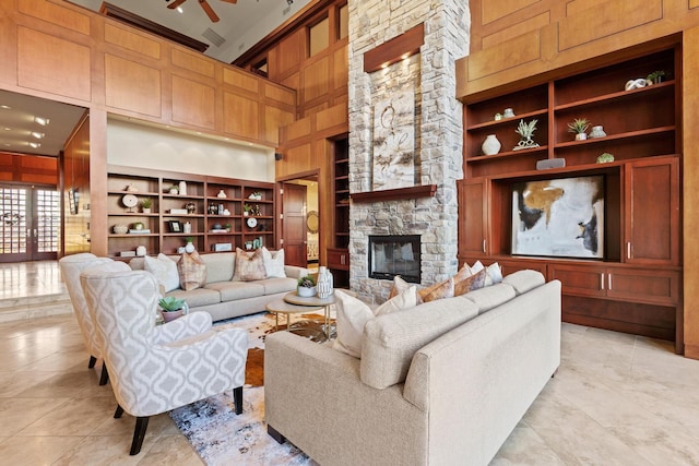 tiled living room with built in shelves, a stone fireplace, wood walls, ceiling fan, and a high ceiling