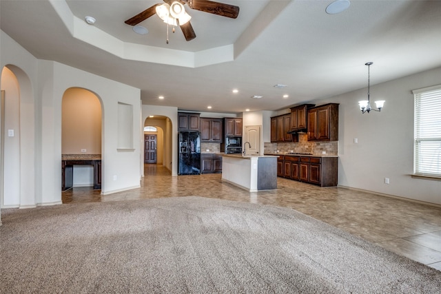 kitchen with ceiling fan with notable chandelier, black appliances, backsplash, a kitchen island with sink, and a tray ceiling