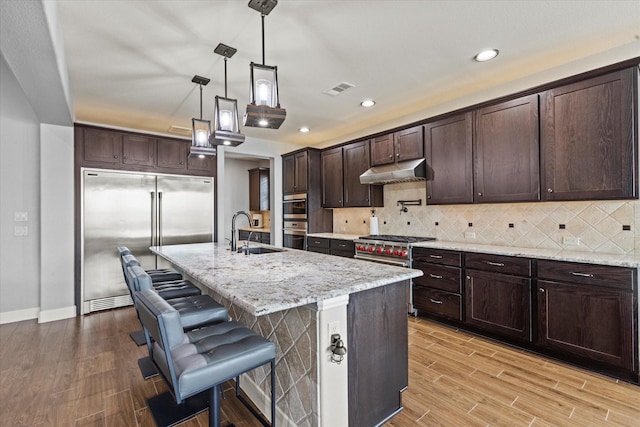 kitchen with sink, high end appliances, decorative light fixtures, an island with sink, and light stone countertops