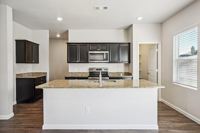 kitchen with light stone counters, dark hardwood / wood-style floors, stainless steel appliances, and an island with sink