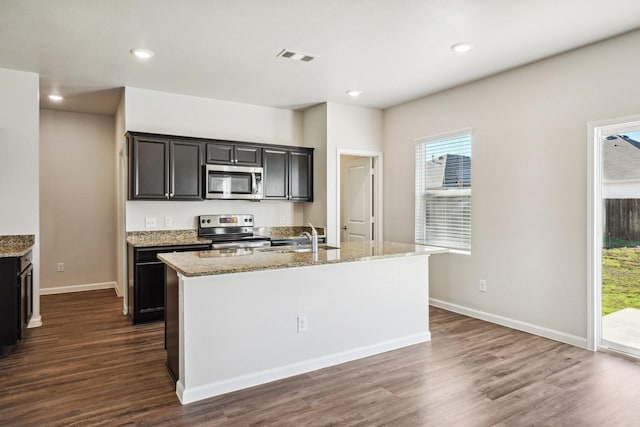 kitchen featuring appliances with stainless steel finishes, an island with sink, sink, light stone counters, and dark wood-type flooring