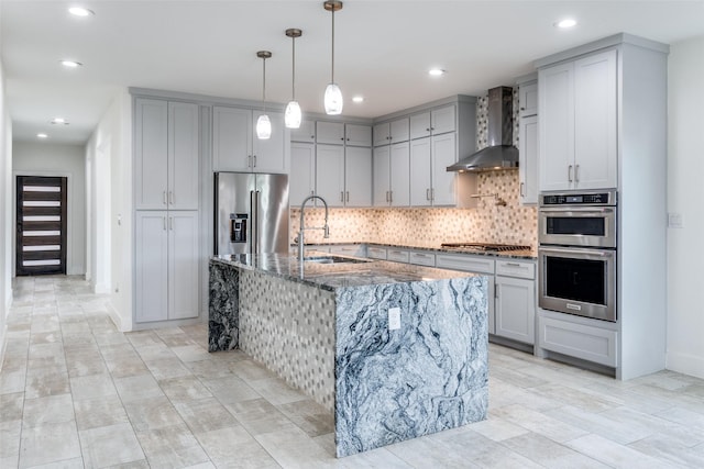 kitchen featuring appliances with stainless steel finishes, a kitchen island with sink, a sink, dark stone countertops, and wall chimney exhaust hood