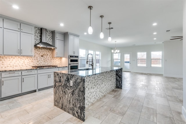 kitchen featuring a kitchen island with sink, a sink, decorative backsplash, wall chimney exhaust hood, and dark stone countertops