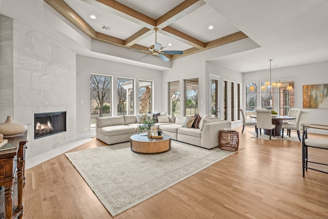 living room featuring coffered ceiling, light wood-type flooring, a tile fireplace, beamed ceiling, and ceiling fan with notable chandelier