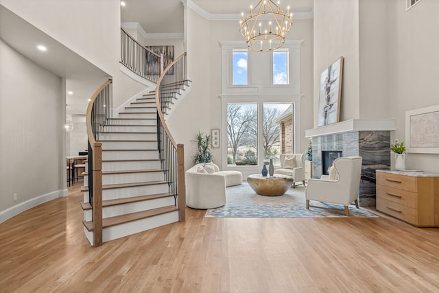 living room featuring crown molding, a towering ceiling, a notable chandelier, a stone fireplace, and light wood-type flooring