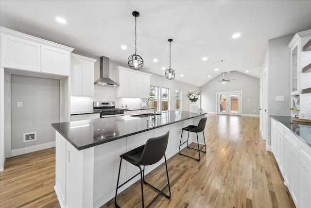 kitchen featuring decorative light fixtures, stainless steel gas stove, white cabinetry, a kitchen island with sink, and wall chimney range hood